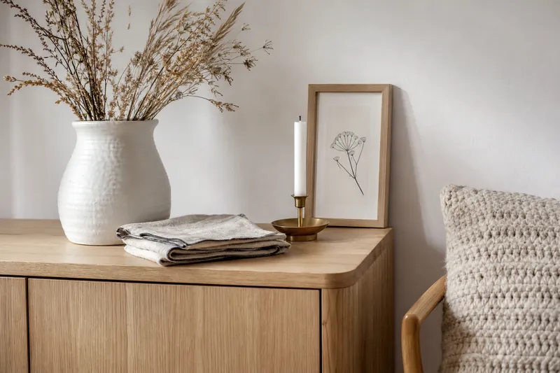 Scandinavian decor details on a light oak sideboard: white ceramic vase with dried grasses, linen napkins, brass candle holder and a framed line print